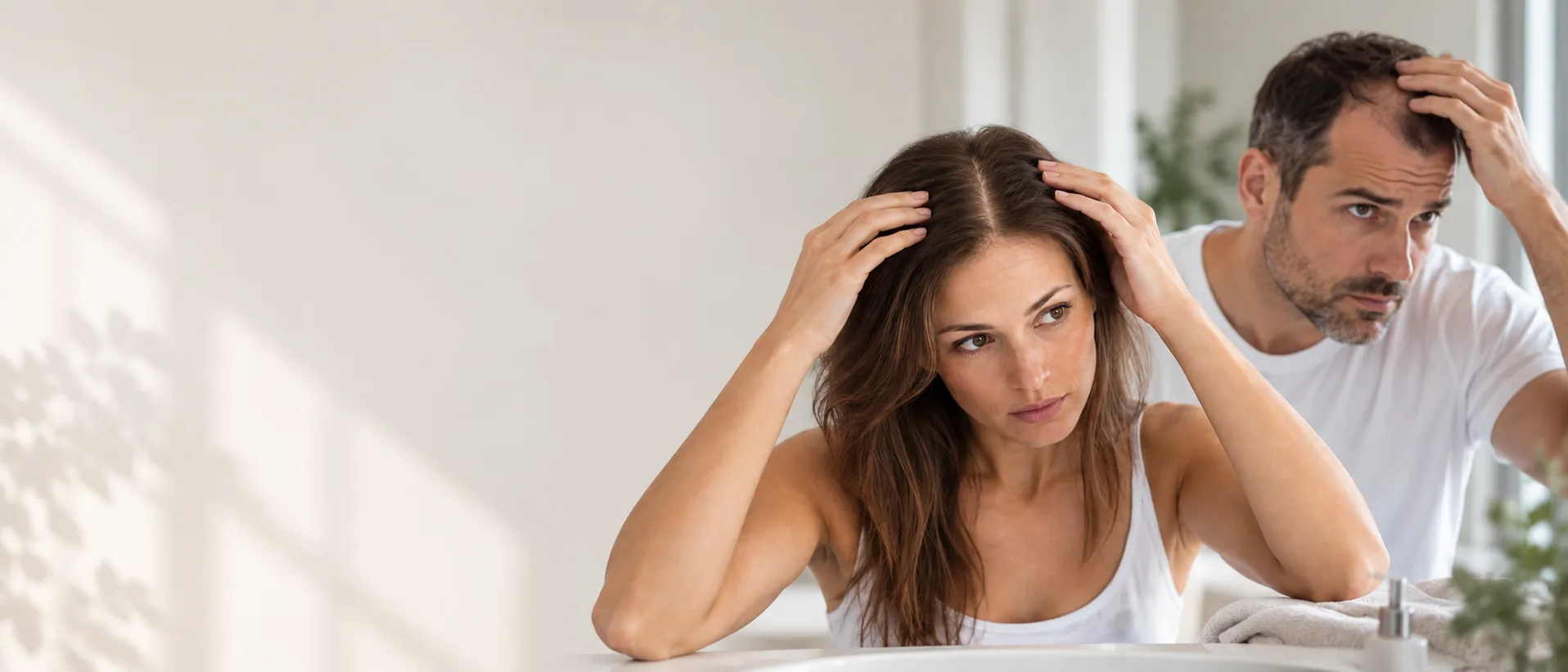 Une femme et un homme devant un miroir de salle de bains, chacun écartant ses cheveux pour observer son cuir chevelu, Clinique esthétique Mozart à Nice
