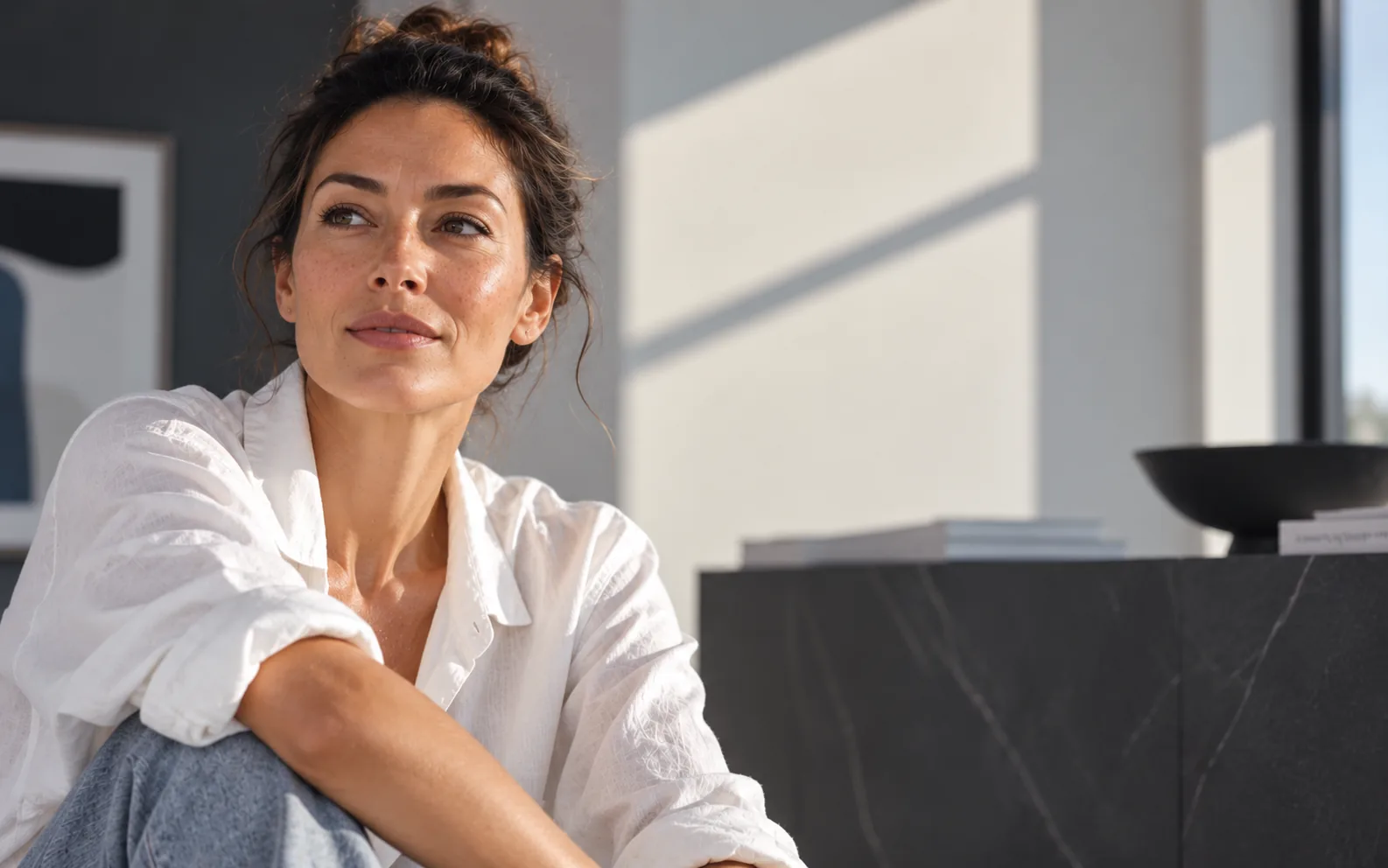 Femme brune en chemise blanche assise dans un intérieur clair, le regard tourné vers la fenêtre